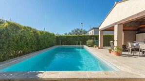 Une piscine rectangulaire bordée de carreaux, entourée d’une haie basse et d’un espace patio aménagé avec du mobilier d’extérieur.