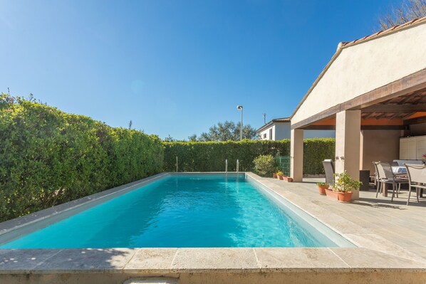 Une piscine rectangulaire bordée de carreaux, entourée d’une haie basse et d’un espace patio aménagé avec du mobilier d’extérieur.