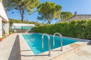 Une piscine rectangulaire avec des échelles en métal, entourée d’une terrasse en pierre, d’une haie et d’arbres en arrière-plan.