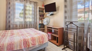 Une chambre à coucher avec un lit, une télévision, une bibliothèque, un ventilateur et une fenêtre avec des rideaux.