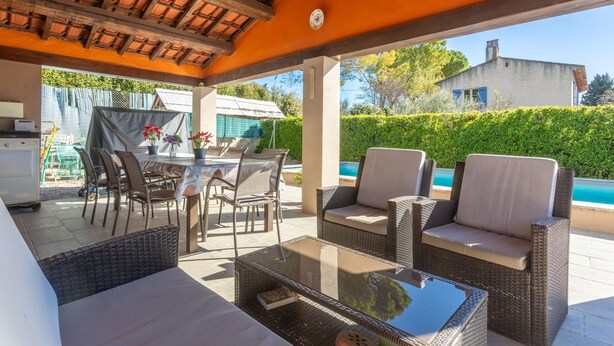 Une terrasse extérieure aménagée avec des meubles en osier, une table en verre et une vue sur une piscine et une maison.