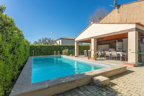 Ein rechteckiger Pool mit Fliesen umgeben von Grünflächen und einem überdachten Außenbereich mit Stühlen und Tischen.