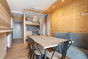 A cozy room with a wooden floor, a dining table with chairs, a sofa, and a kitchen area with cabinets and a countertop. - Cozy 52 m² Apartment – Ski-In/Ski-Out Access, Balcony & Wi-Fi in Alpe d'Huez (Huez)