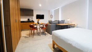 Moderni hotellihuone, jossa on sänky, sohva, ruokapöytä tuoleineen, keittiö sekä seinälle asennettu televisio.