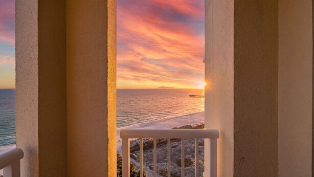 Ein Balkon mit Blick auf das Meer bei Sonnenuntergang, ein Steg in der Ferne und eine felsige Küstenlinie.