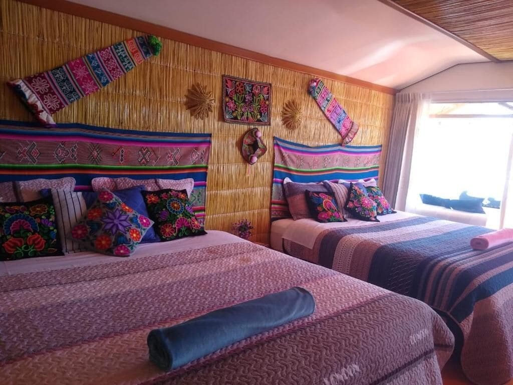A room with two beds, each with colorful bedding and pillows, a bamboo wall, and a window with curtains.