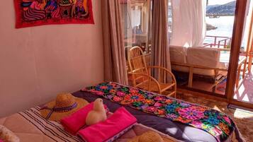 A room with a bed, a colorful woven blanket, a straw hat, and a view of the sea through a glass door.