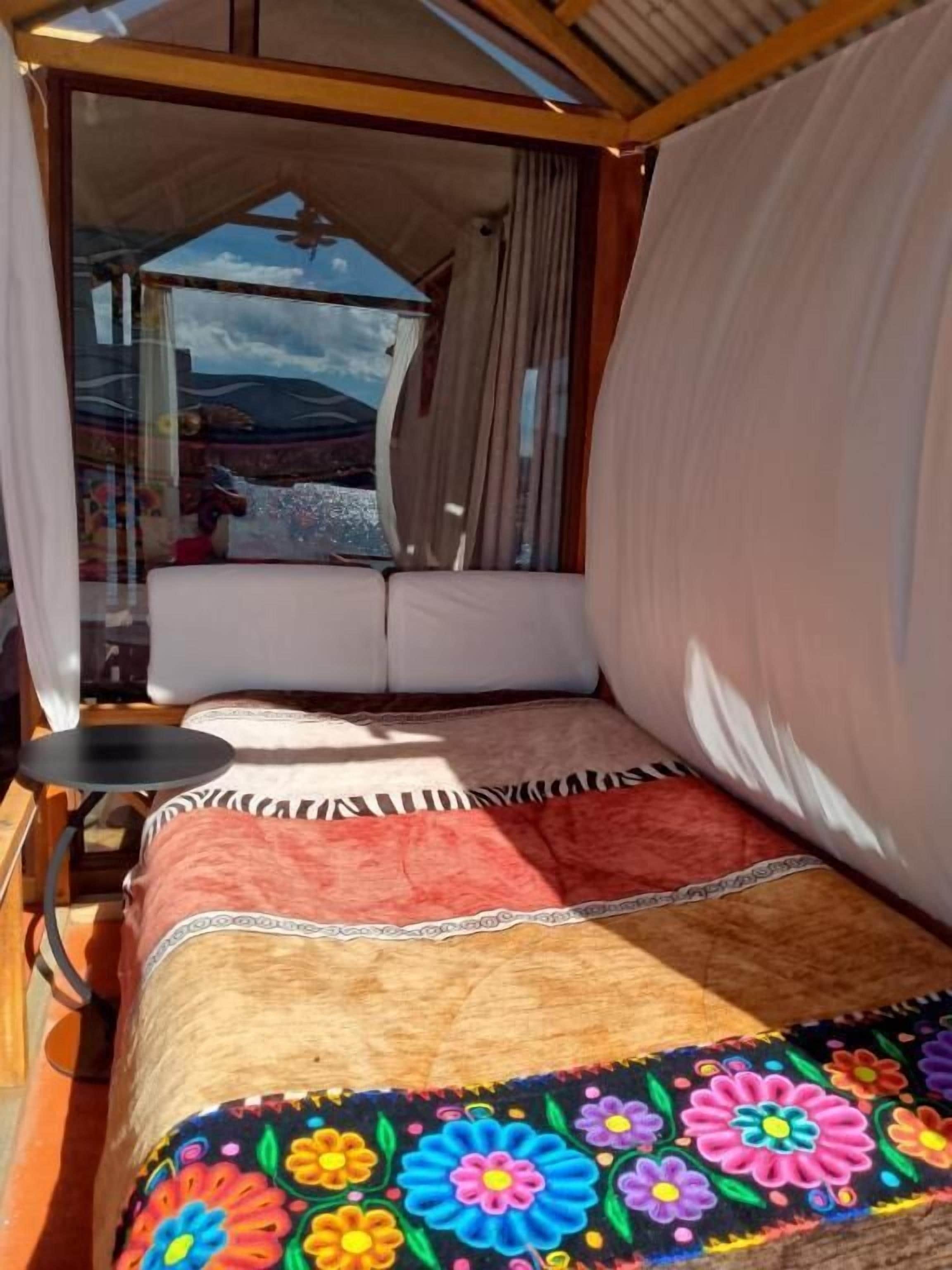 A bed with a colorful floral bedspread, a small table, and a view of a mountainous landscape through a window.
