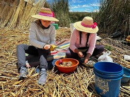 Dos personas sentadas sobre paja, realizando una actividad con cuencos y baldes, rodeadas de vegetación natural.