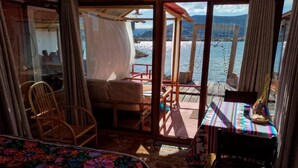 A room with a bed, a chair, a table with a colorful tablecloth, and a view of a body of water through a glass door. - Uros TITIKAKA the Best lodge  (Puno)
