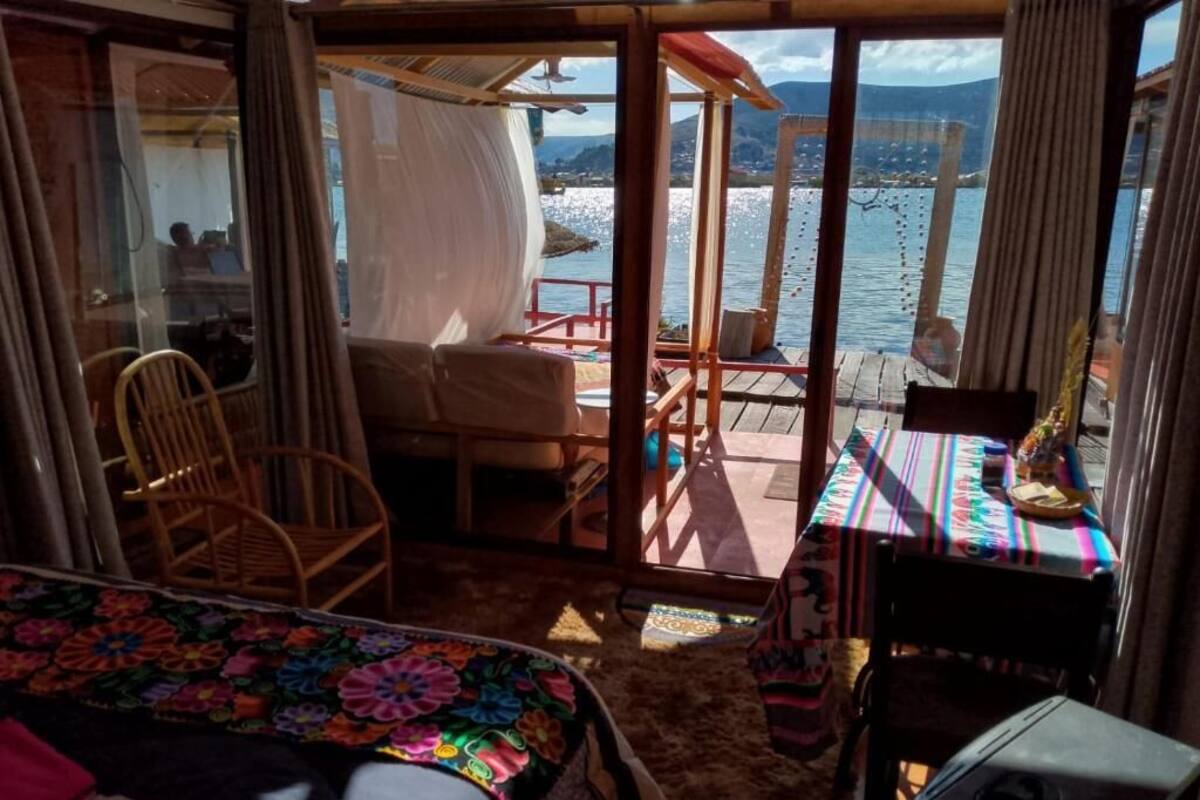 Una habitación con cama, una silla, una mesa con mantel colorido y vista a un cuerpo de agua a través de una puerta de cristal.