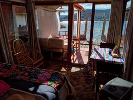 Una habitación con cama, una silla, una mesa con mantel colorido y vista a un cuerpo de agua a través de una puerta de cristal.