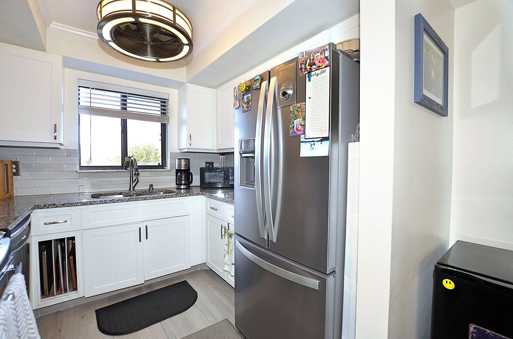 A modern kitchen with white cabinets, a stainless steel refrigerator, a coffee maker, and a window with blinds.