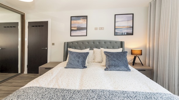 A hotel room with a large bed, two nightstands, a lamp, and two framed pictures on the wall. - Brand New Flat with AC 5-Min Walk to Town Centre - Pass the Keys (Reading)