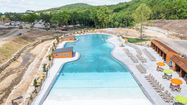 Uma piscina grande com design ondulado, cercada por cadeiras de descanso e guarda-sóis, com colinas e árvores ao fundo.