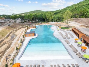 Une grande piscine à la forme ondulée, entourée de chaises longues et de parasols, avec en arrière-plan des collines et des arbres.