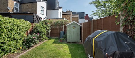 A residential backyard with a lawn, a wooden shed, and a barbecue grill covered with a protective sheet.