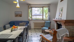 Een eetgedeelte met een tafel, stoelen, een blauwe bank, een open haard en een raam, buiten staat een parasol.