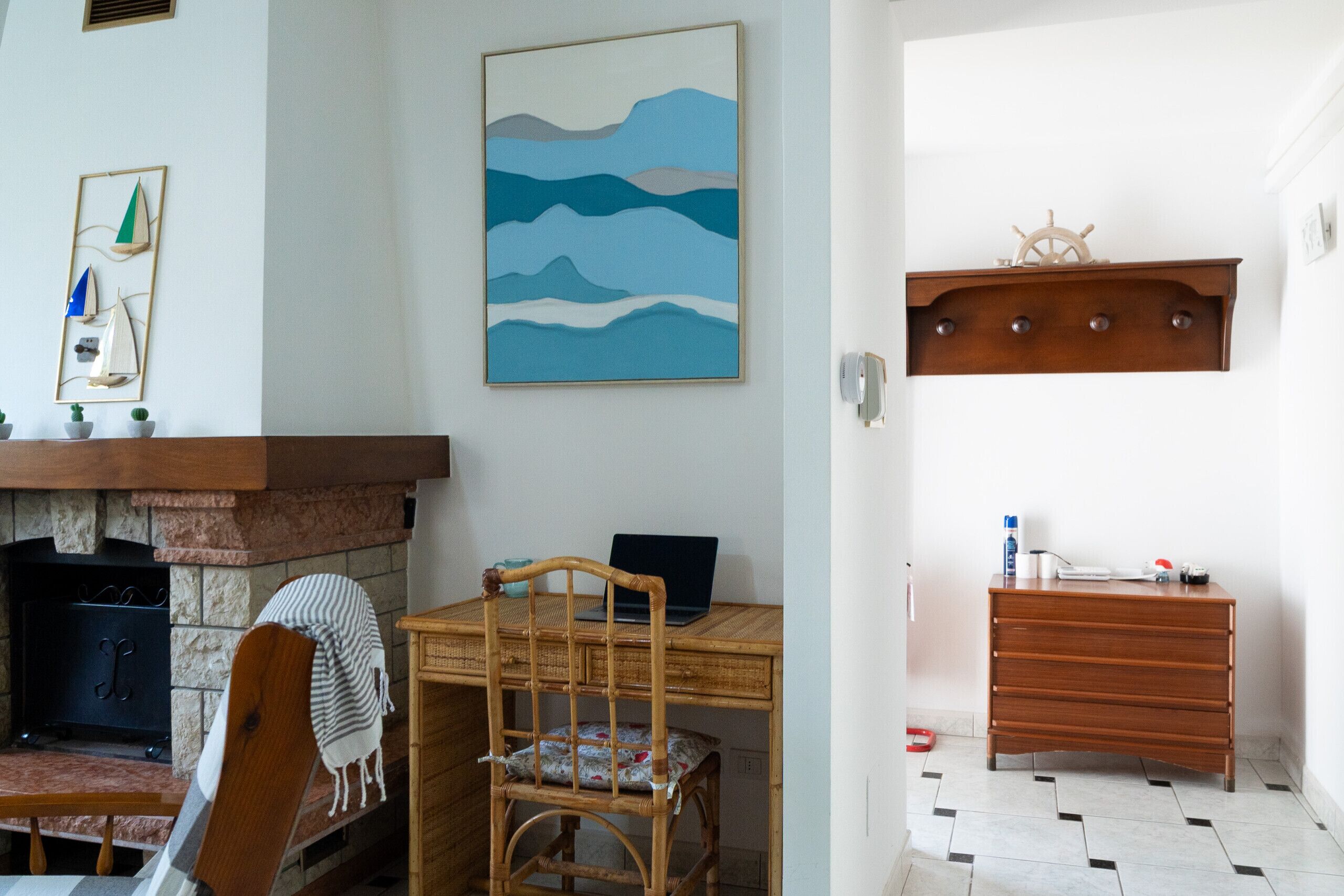 Une pièce avec une cheminée, un ordinateur portable posé sur un bureau en bois, une chaise, une étagère fixée au mur ornée d’un gouvernail de bateau, et un sol carrelé.