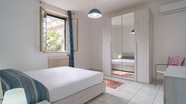 Een slaapkamer met een bed, nachtkastje, lamp, raam met gordijnen, kast en stoel.