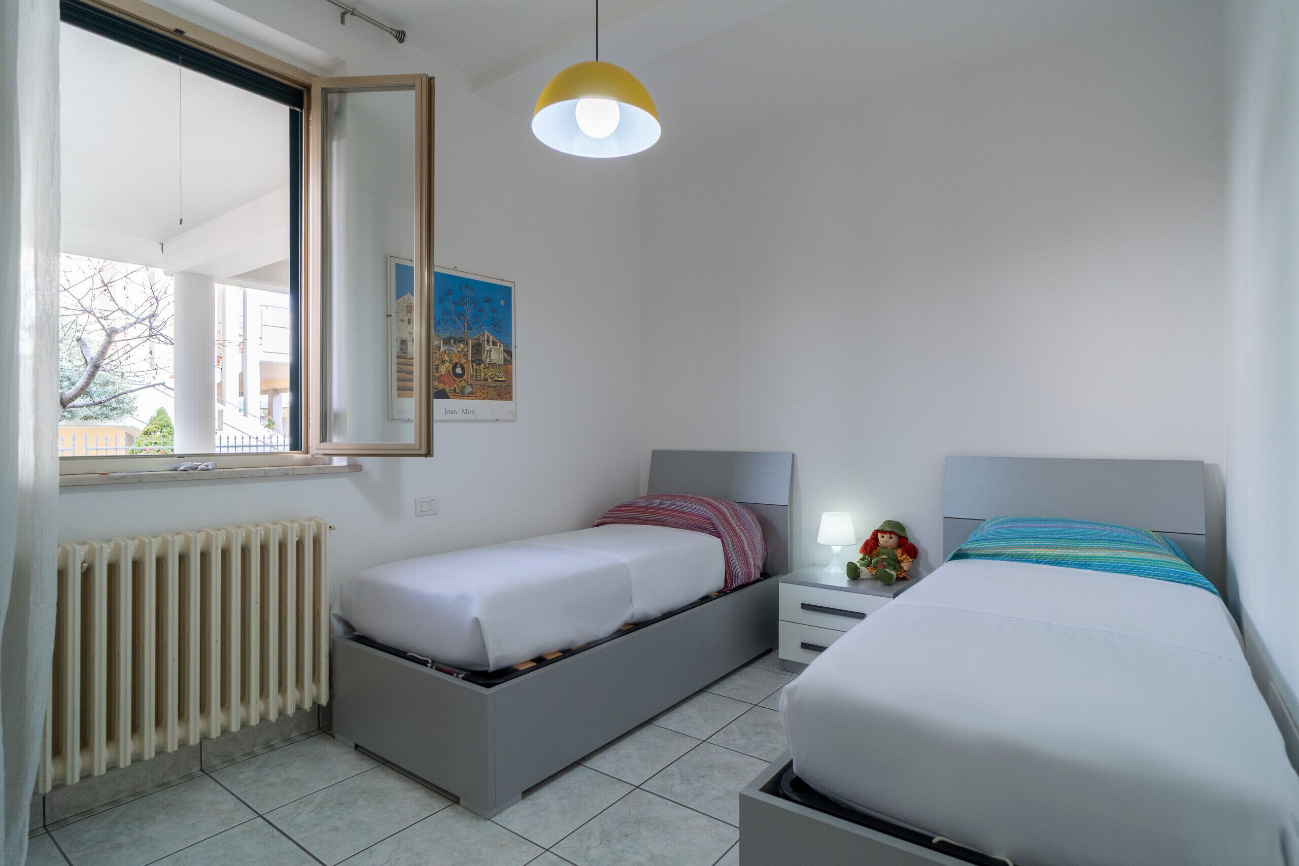 A bedroom with two beds, a window, a radiator, and a hanging light fixture.