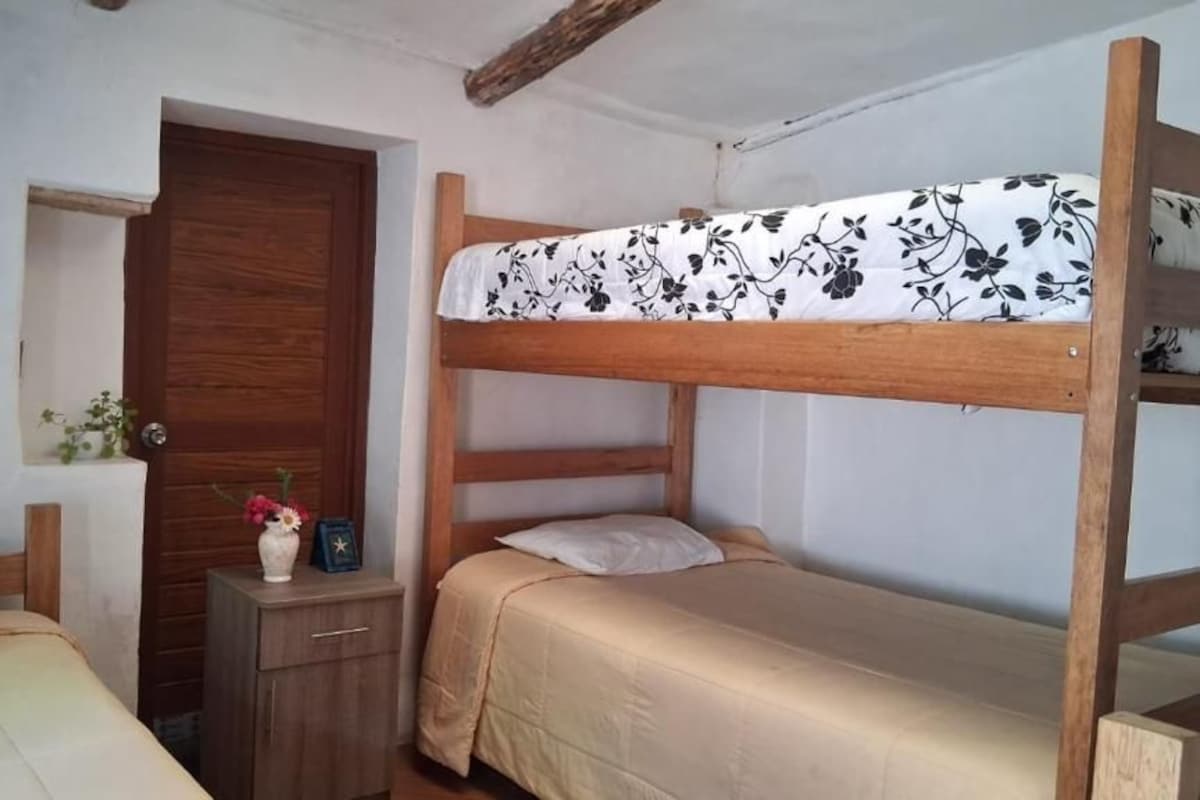 Une pièce avec deux lits superposés, une porte en bois, une petite table de chevet avec un vase et un cadre photo, et une étagère fixée au mur.