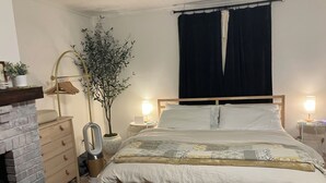 Et soveværelse med seng, sengeborde, en kommode, et spejl, en potteplante og et lille bord med lampe.