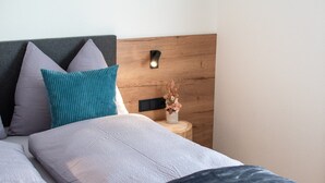 Un lit bien fait, avec une tête de lit, des oreillers, une table de chevet ornée d’un vase de fleurs et une lampe fixée au mur.
