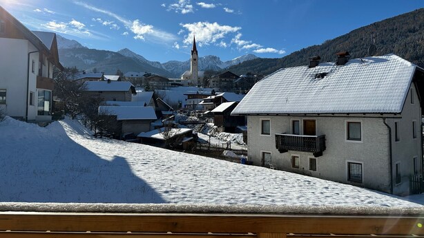 Un village enneigé, avec des bâtiments traditionnels, un clocher d’église et des montagnes en arrière-plan.