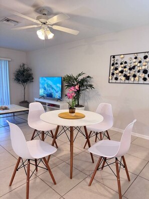 Comedor con una mesa redonda, cuatro sillas blancas, un ventilador de techo, un televisor y una obra de arte en la pared.