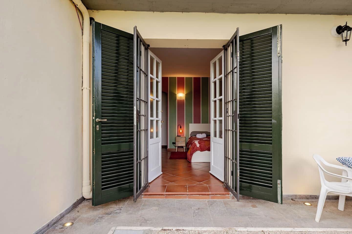 A hotel entrance with open green shutters, a tiled floor, a bed with a red blanket, and a white chair.