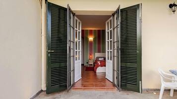 A hotel entrance with open green shutters, a tiled floor, a bed with a red blanket, and a white chair.