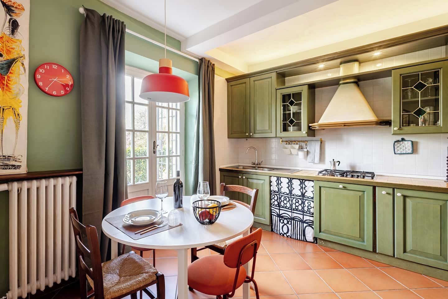 A kitchen with green cabinets, a white countertop, a red clock, and a dining area with a round table and chairs.