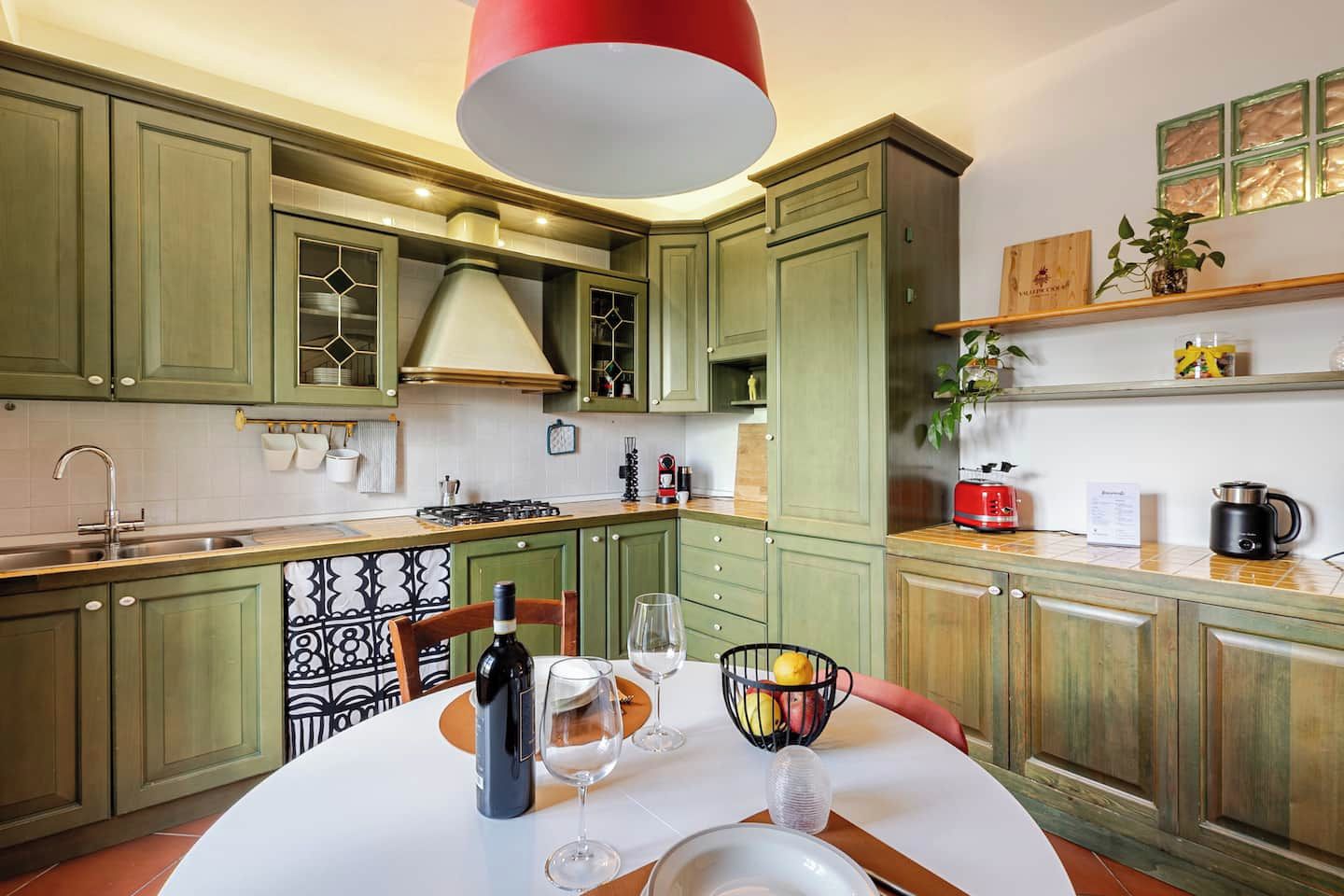 A kitchen with green cabinets, a white countertop, a red hanging light, and a dining table set with wine glasses and a bowl of fruit.