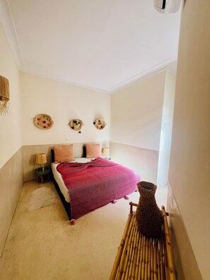 Een slaapkamer met een bed, kussens, een lamp, een decoratief object aan de muur en een rotan stoel.