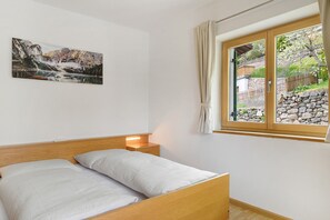 Ein Schlafzimmer mit einem hölzernen Bett, einem Nachttisch, einer Wandlampe, einem Fenster mit Vorhängen und einem Gemälde einer Berglandschaft.