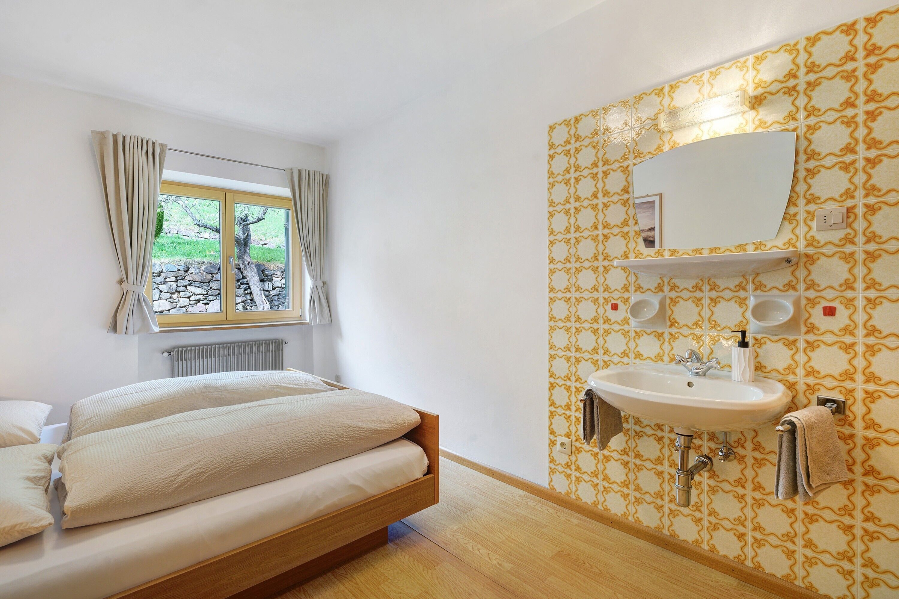 Een slaapkamer met een bed, een raam met gordijnen, een badkamer met een wastafel en spiegel, en behang met een patroon.