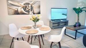 Moderni olohuone, jossa on ruokapöytä, valkoiset tuolit, litteänäyttöinen televisio ja taulu seinällä.