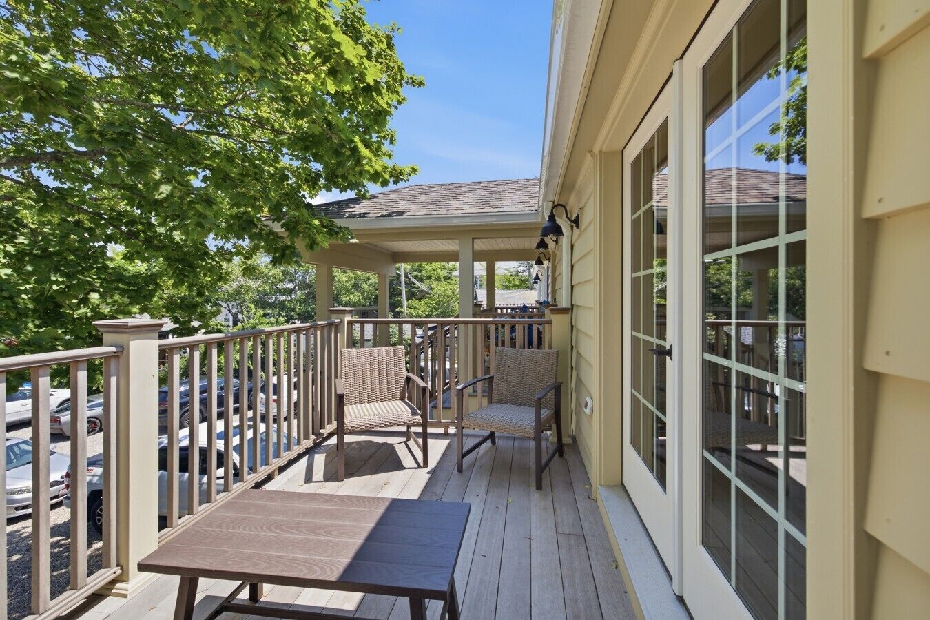 A covered porch with a table and chairs, a railing, and a view of parked cars and trees.