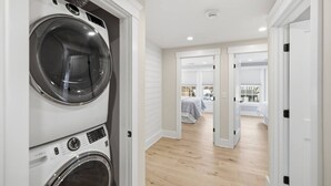 Lavanderia con lavatrici e asciugatrici affiancate, pavimento in legno e un corridoio che porta a una camera da letto con un letto e una finestra.