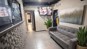 Una zona de recepció amb un taulell d'entrada, un sofà gris, un televisor penjat a la paret i un extintor.