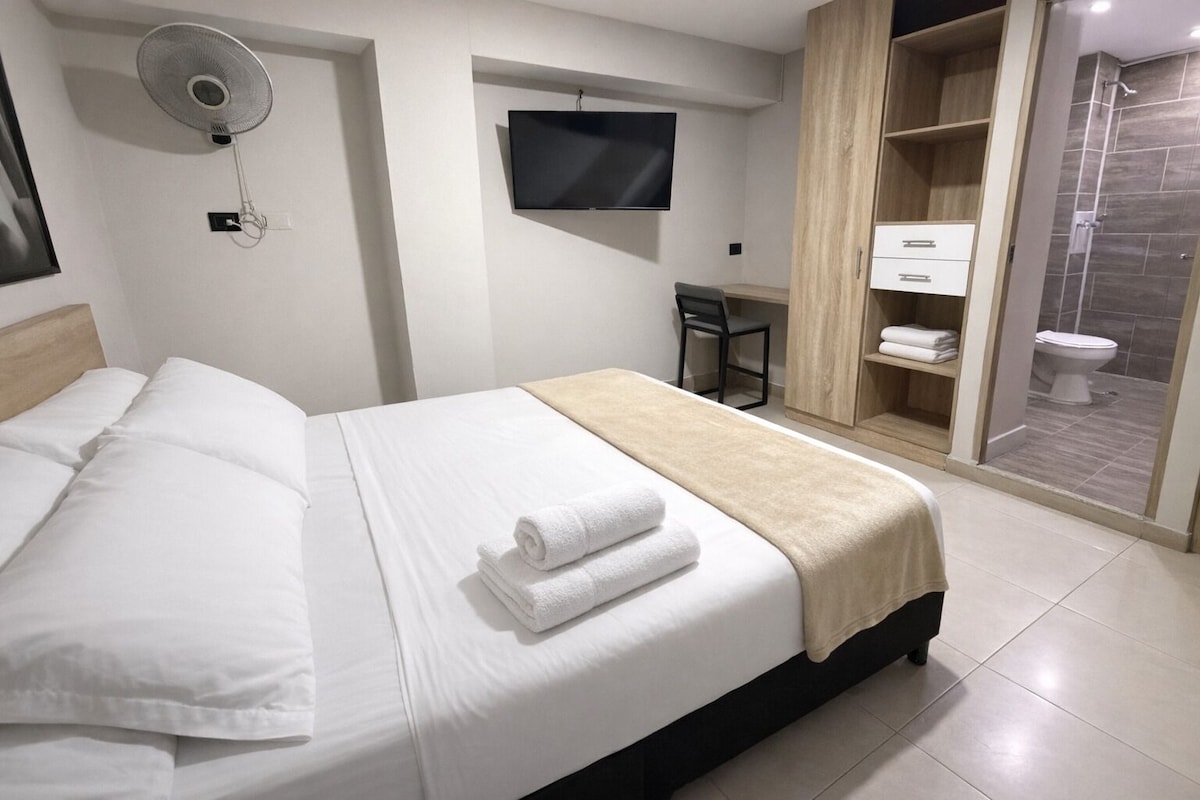 A hotel room with a bed, a TV, a desk, a chair, a shower area, and a toilet.