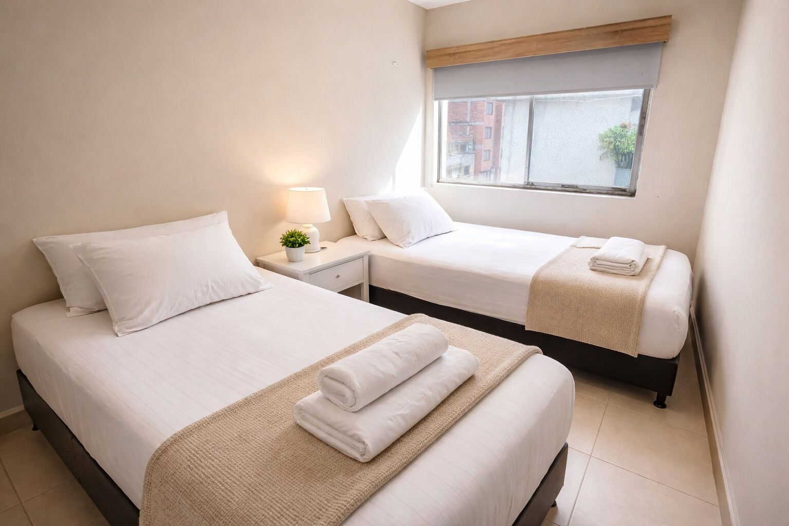 Una camera d'albergo con due letti, un comodino con una lampada, una finestra vista edifici e una piccola pianta sul comodino.