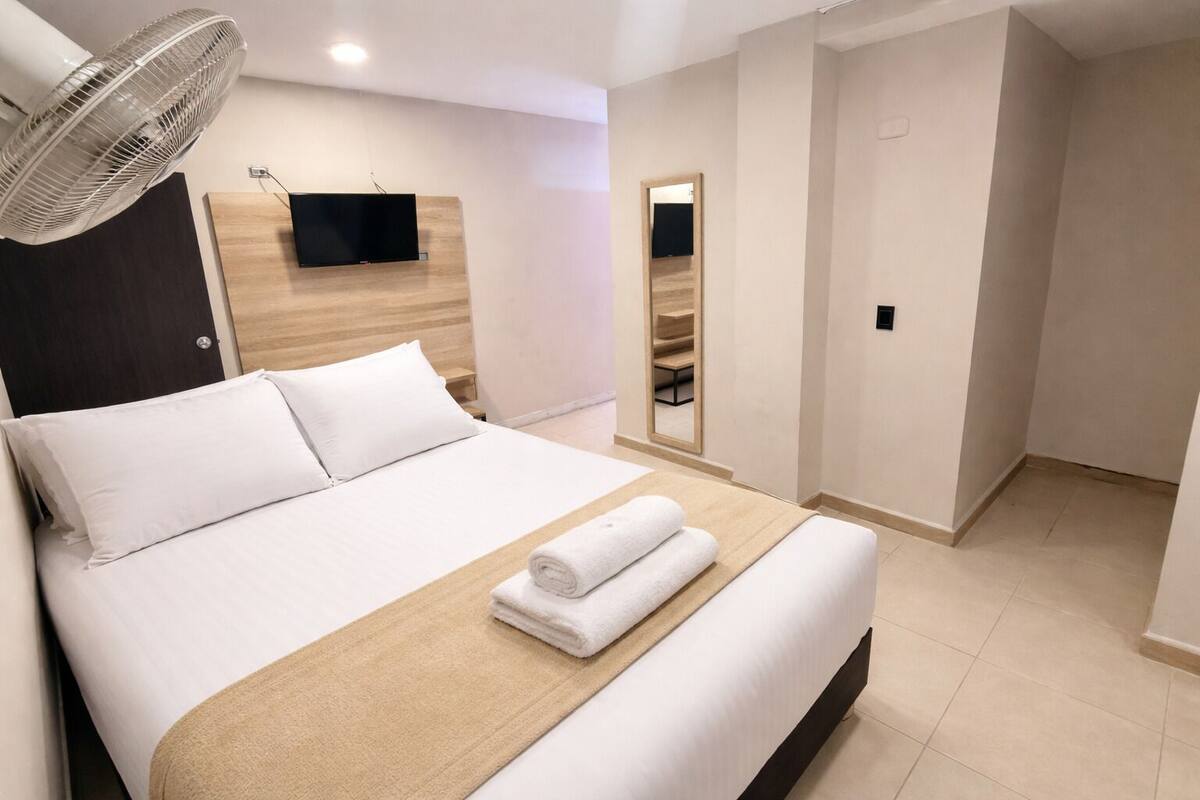 Una habitació d'hotel amb un llit, un televisor penjat a la paret, un ventilador de peu i un mirall.