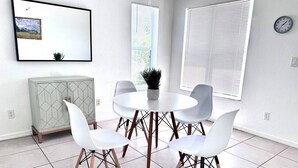 A modern dining area with a round table, four white chairs, a geometric cabinet, and a painting on the wall. - SWFL: Cape Coral Relax - Entire Home! (Cape Coral)