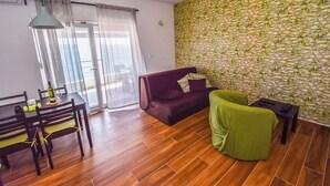 A living room with a wooden floor, a purple sofa, a green chair, a dining table with chairs, and a wall with a leafy pattern. - Marli Beach Apartments Jesenice Marli 1 (Dugi Rat)