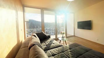 Ein modernes Wohnzimmer mit einer großen Couch, einer Glastür, die auf einen Balkon führt, einem runden Tisch mit einer Vase und einem an der Wand befestigten Fernseher.