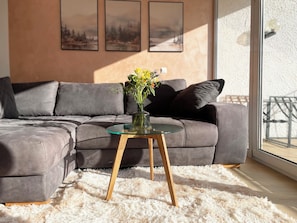 Ein modernes Wohnzimmer mit einem grauen Ecksofa, einem Glastisch und einem flauschigen weißen Teppich.