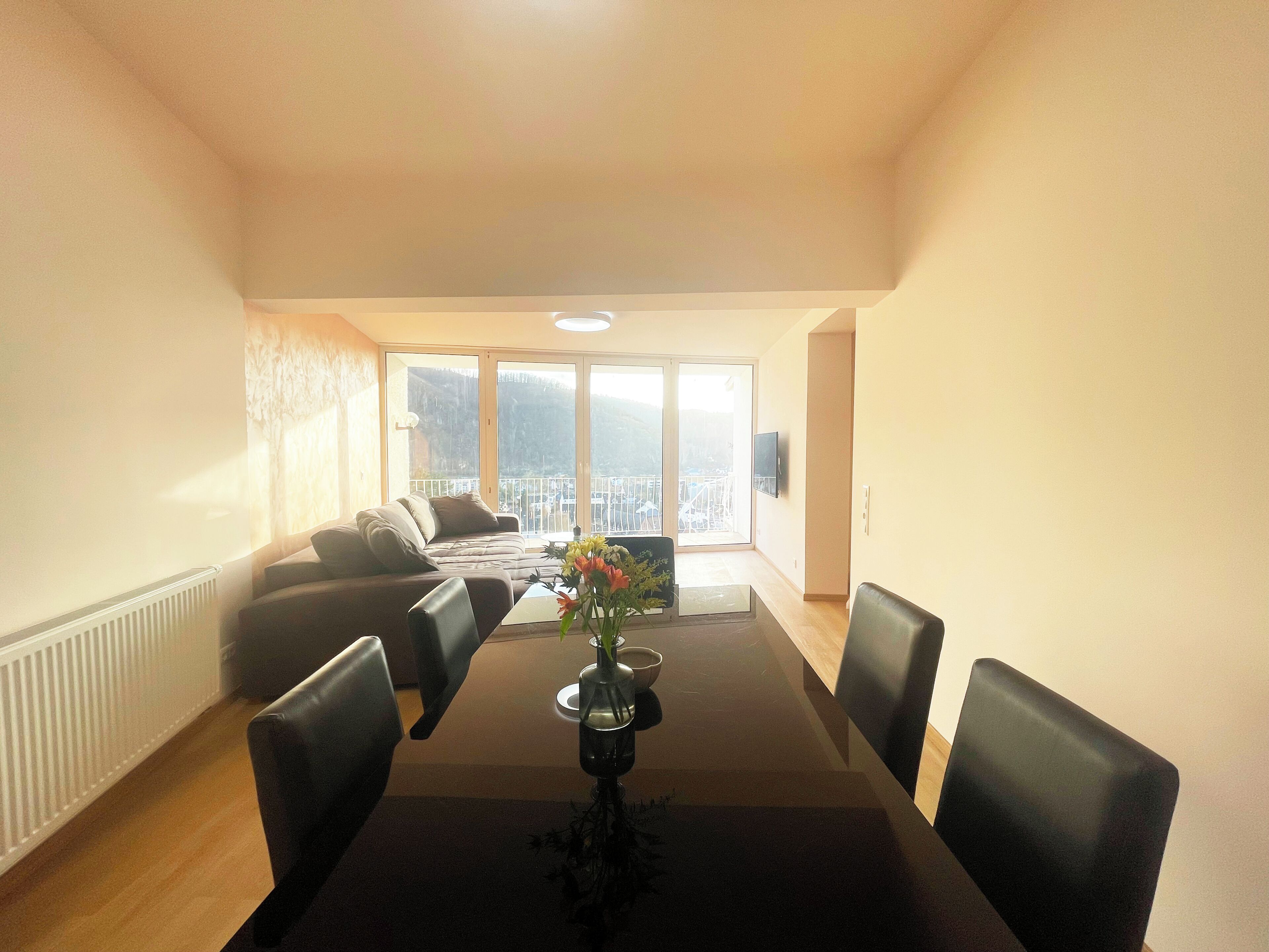A dining area with a table, chairs, a vase with flowers, a sofa, and a view of a cityscape through large windows.