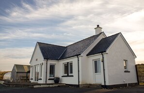 A white house with a dark roof, a chimney, and a front porch. - Solas Alainn -  a detached house that sleeps 6 guests  in 3 bedrooms (Staffin)